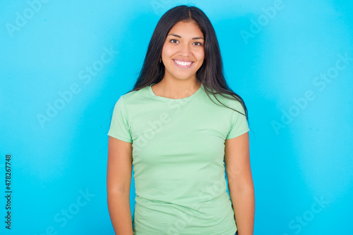 Young hispanic girl wearing green T-shirt over blue background with nice beaming smile pleased expression. Positive emotions concept © Roquillo