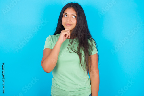 Thoughtful Young hispanic girl wearing green T-shirt over blue background holds chin and looks away pensively makes up great plan