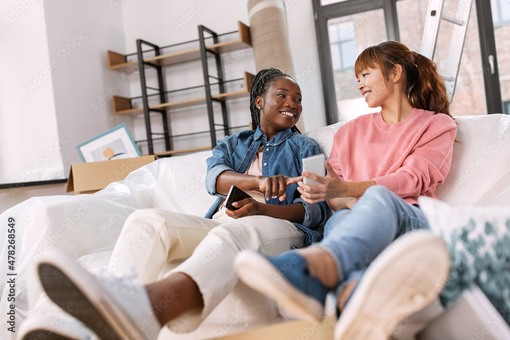 moving, people and real estate concept - happy smiling women with smartphones and boxes at new home moving, people and real estate concept - happy smiling women with smartphones and boxes at new home