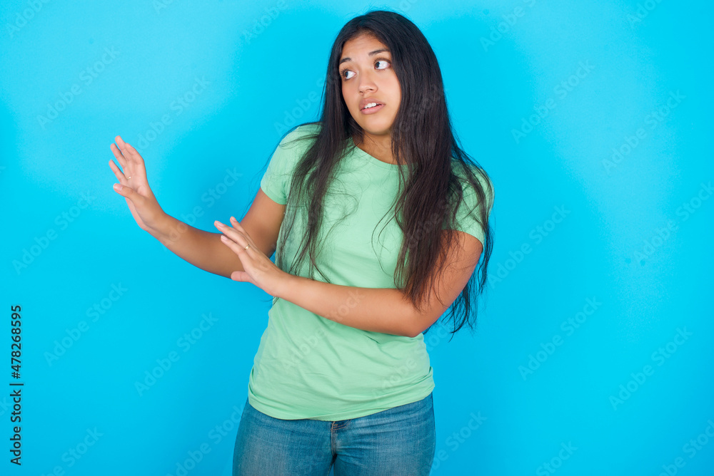 Displeased Young hispanic girl wearing green T-shirt over blue background keeps hands towards empty space and asks not come closer sees something unpleasant Displeased Young hispanic girl wearing green T-shirt over blue background keeps hands towards empty space and asks not come closer sees something unpleasant