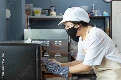 Happy young Asian woman with protective face mask, apron is smiling while using oven to bake homemade cake or bread in home kitchen, learning new skills when home quarantine, COVID-19 or Coronavirus.