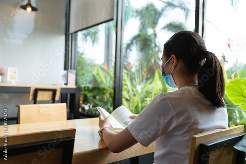 back of happy young Asian woman with medical protective face mask, white casual t-shirt is smiling, reading book in cafe, coffee shop with little cute green tree with social distancing in COVID19