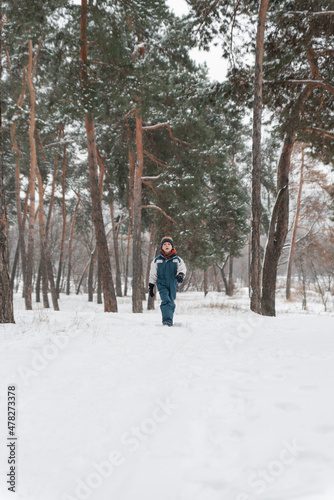 Boy in winter jumpsuit and multi-colored hat comes out snow-covered forest. Walk against of beautiful winter landscape