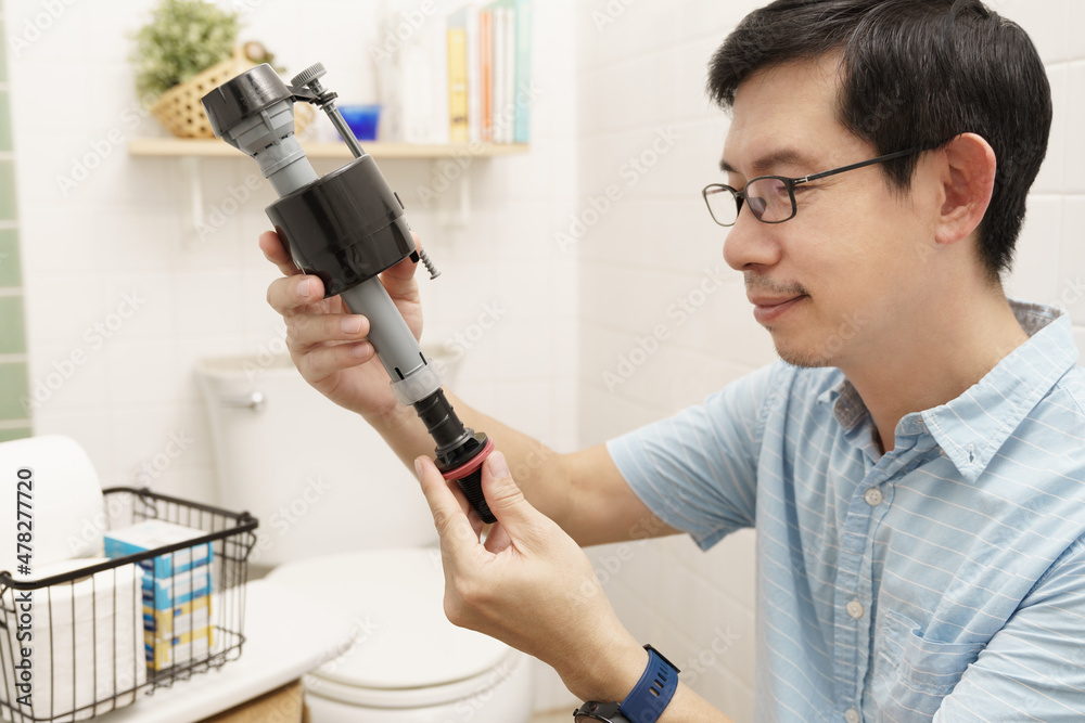Happy middle aged and handsome Asian man check new fill valve before fix and replace broken valve in toilet tank in clean white bathroom. DIY plumbing, home maintenance, handyman and house improvement Happy middle aged and handsome Asian man check new fill valve before fix and replace broken valve in toilet tank in clean white bathroom. DIY plumbing, home maintenance, handyman and house improvement