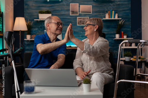 Quadro em tela Caucasian old people highfive while sitting on sofa using laptop at home