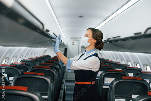 In protective gloves and mask. Young stewardess on the work in the passanger airplane