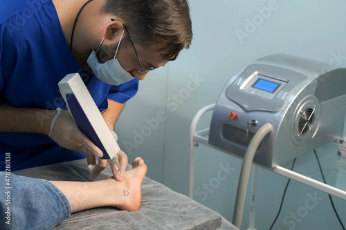 Laser removing of tattoo with words loser  lover and heart on woman s foot in red and black colours  closeup hands of doctor in gloves. Romantic tattoo symbol of youth love and disappointment in life.