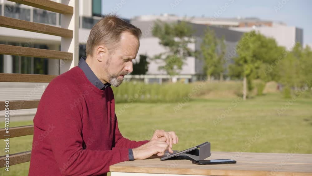 A middle-aged handsome Caucasian man works on a tablet as he sits at a table in a park in an urban area - office buildings in the blurry background - closeup
