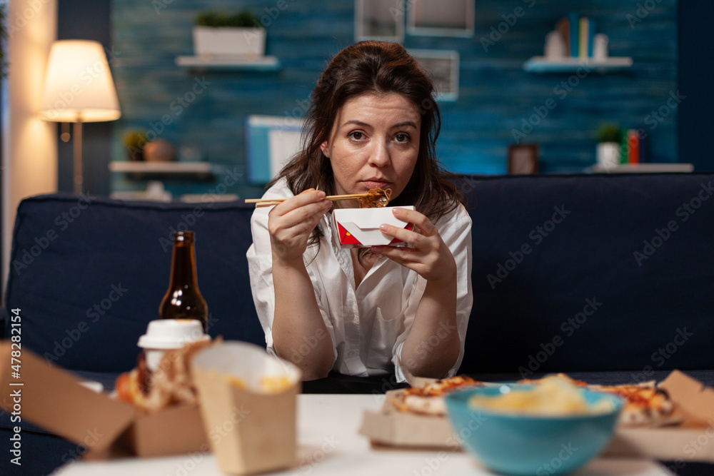 Woman eating takeout asian noodles with chopsticks on living room sofa while watching news on television. Office worker sitting on couch in the evening enjoying chinese takeaway ramen box. Woman eating takeout asian noodles with chopsticks on living room sofa while watching news on television. Office worker sitting on couch in the evening enjoying chinese takeaway ramen box.