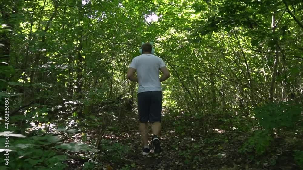 a young man in a white meek and gray shorts with headphones runs through the forest on a summer morning. the bright sun shines, hard shadows. a young man in a white meek and gray shorts with headphones runs through the forest on a summer morning. the bright sun shines, hard shadows.