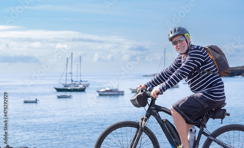 Happy senior man cyclist wearing sunglasses, backpack and helmet riding in front of the sea with his bicycle enjoying outdoors and freedom. Horizon over the water, blue sky