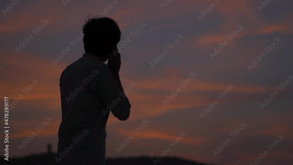 Silhouette shot of an Indian man talking on the mobile phone while looking at the orange clouds during the sunset. Happy teenager talking on the mobile phone. Teen talking with his friends on phone. Silhouette shot of an Indian man talking on the mobile phone while looking at the orange clouds during the sunset. Happy teenager talking on the mobile phone. Teen talking with his friends on phone.