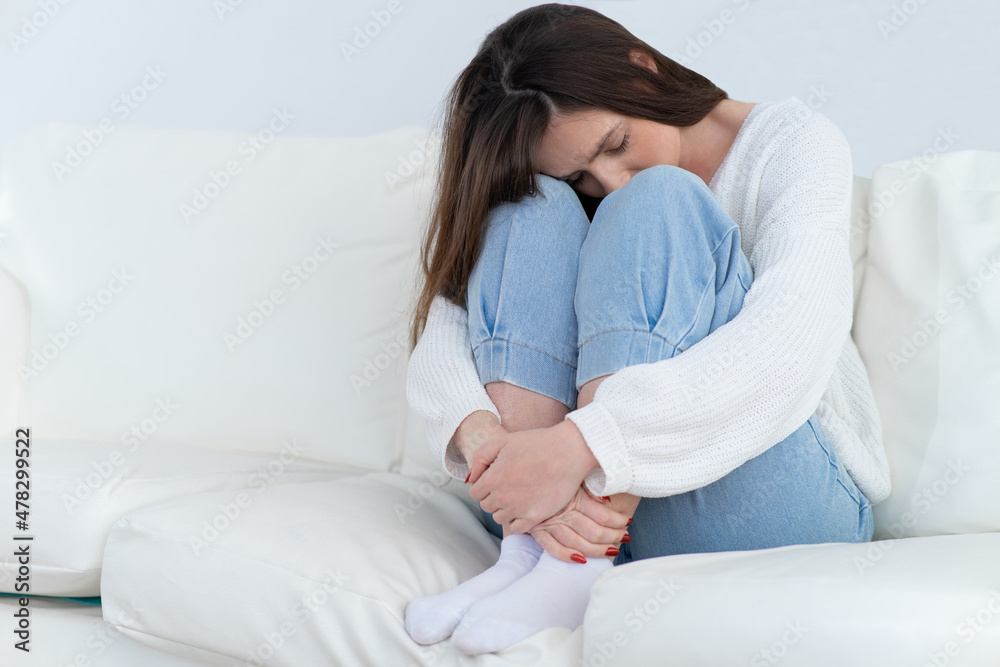 Young brunette woman is depressed. Psychological help is needed. Sitting on the sofa with his head on his knees. White sofa and walls. Young brunette woman is depressed. Psychological help is needed. Sitting on the sofa with his head on his knees. White sofa and walls.