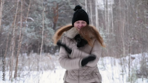 Red-haired girl jumping, laughing and dancing in winter forest in warm clothes: black hat and light jacket, 4k. Blurred forest background