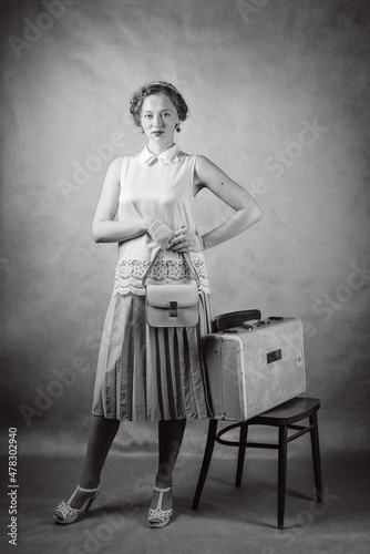 Young woman, dressed in retro style, with a handbag in her hands and a suitcase on a chair. Studio black and white portrait