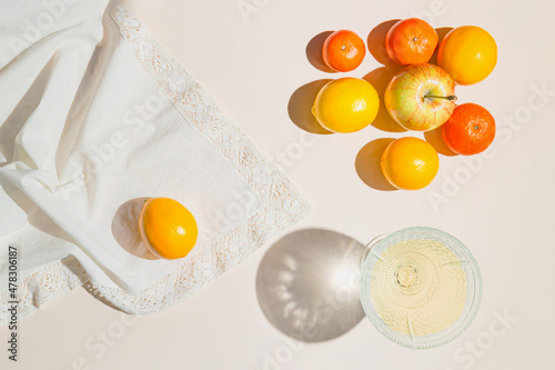 Creative composition made of glass with fruit juice, lemon, mandarin and apples on pastel beige sunlit background with white cloth and shadows. Summer and refreshment concept. Harvest theme