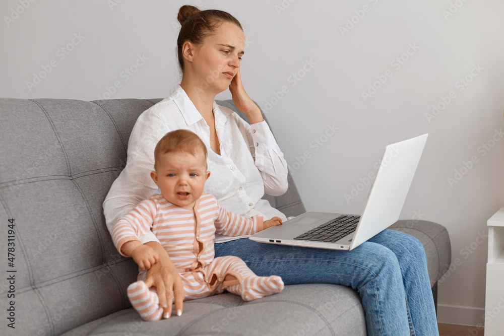 Indoor shot of young adult dark haired woman with laptop on the sofa with her baby, doing remote work, feeling stress, being tired to work and taking care of her child.
