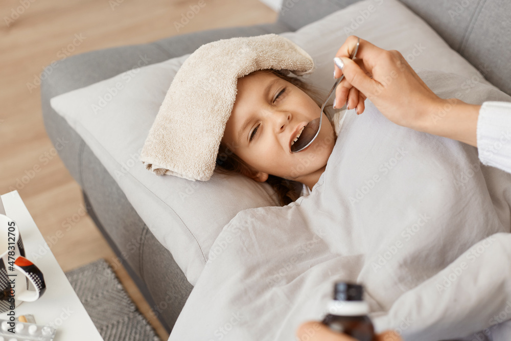 Horizontal shot of little cute sick girl with towel on her forehead lying on sofa, mother giving her spoon with syrup, treating her child with influenza and high temperature. Horizontal shot of little cute sick girl with towel on her forehead lying on sofa, mother giving her spoon with syrup, treating her child with influenza and high temperature.