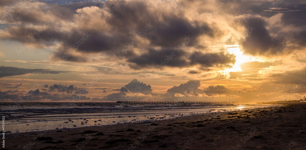 Sunset over a beach, with mesmerising clouds and sea foam left on the beach after the receding waves. A pier can be seen in the distance.