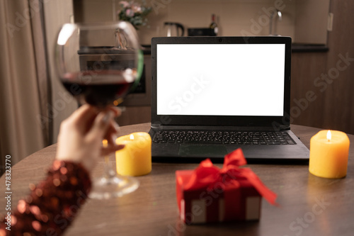 A blank laptop screen stands on the table. A woman's hand raises a glass of red wine. concept of dating online at a distance.