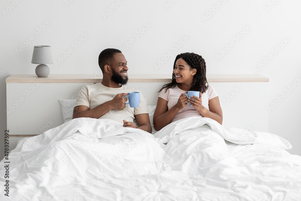 Smiling happy millennial african american man and wife sit on bed in white bedroom interior with cups of coffee Smiling happy millennial african american man and wife sit on bed in white bedroom interior with cups of coffee