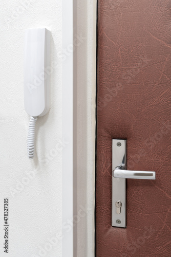 Simple white intercom or house phone attached to a wall next to a closed door with metallic handle. Home safety and communication. © Petr