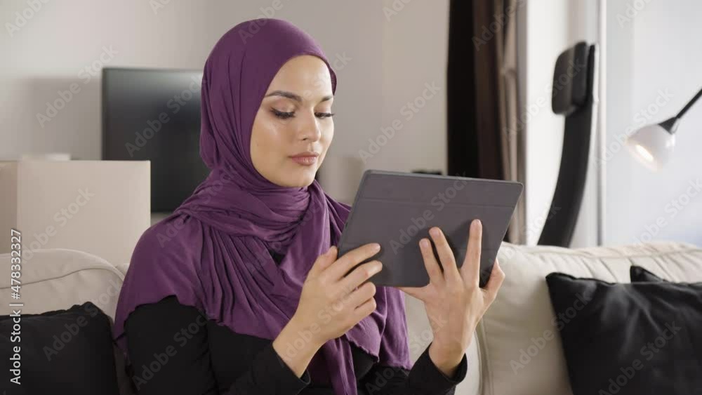 A young beautiful Muslim woman works on a tablet with a smile as she sits on a sofa in an apartment A young beautiful Muslim woman works on a tablet with a smile as she sits on a sofa in an apartment