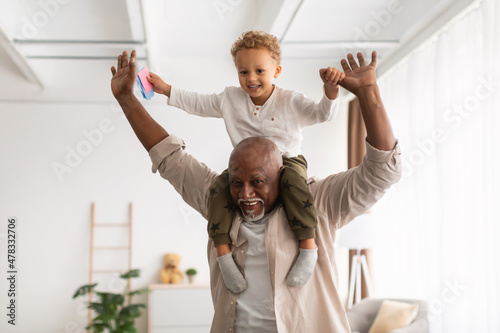 Cheerful Black Grandpa Carrying Little Grandson On Shoulders Playing Indoor photo