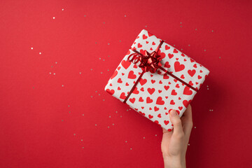 First person top view photo of st valentine's day decor female hand giving giftbox in white wrapping paper with heart pattern red star bow and sequins on isolated red background with empty space
