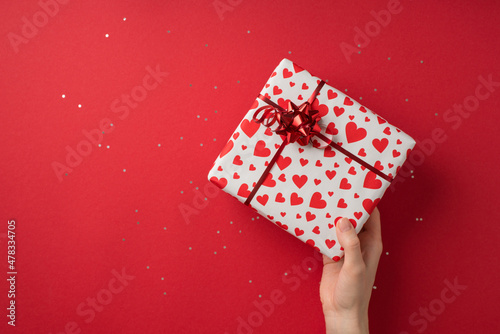 First person top view photo of st valentine's day decor female hand giving giftbox in white wrapping paper with heart pattern red star bow and sequins on isolated red background with empty space photo