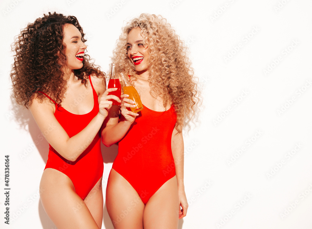 Two beautiful sexy smiling hipster women in red summer swimwear bathing suits.Trendy models with afro curls hairstyle having fun in studio.Hot female isolated on white.Drinking lemonade from bottle
