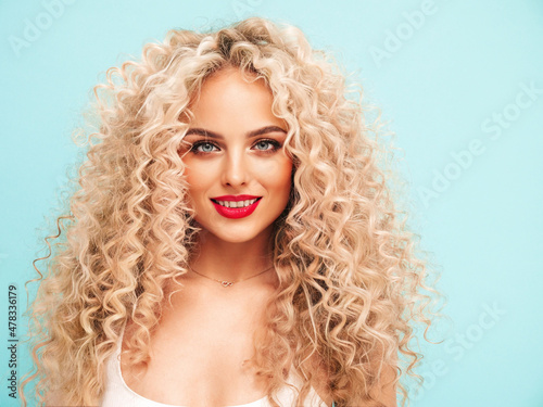 Closeup portrait of young beautiful smiling blond hipster female in trendy summer clothes. Sexy carefree woman posing near blue wall with afro curls hairstyle. Positive model having fun