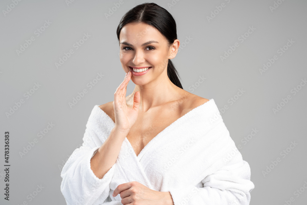Beautiful woman in white bathrobe touching her face with the hand isolated on gray background. Perfect morning concept. Brunette female with long shinning hair in bathroom Beautiful woman in white bathrobe touching her face with the hand isolated on gray background. Perfect morning concept. Brunette female with long shinning hair in bathroom