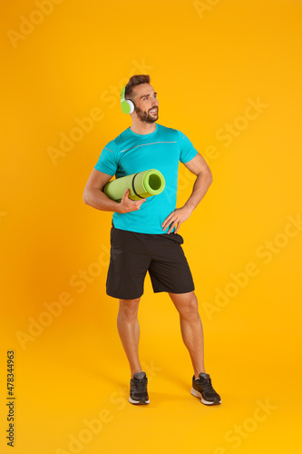 Handsome man with yoga mat and headphones on yellow background