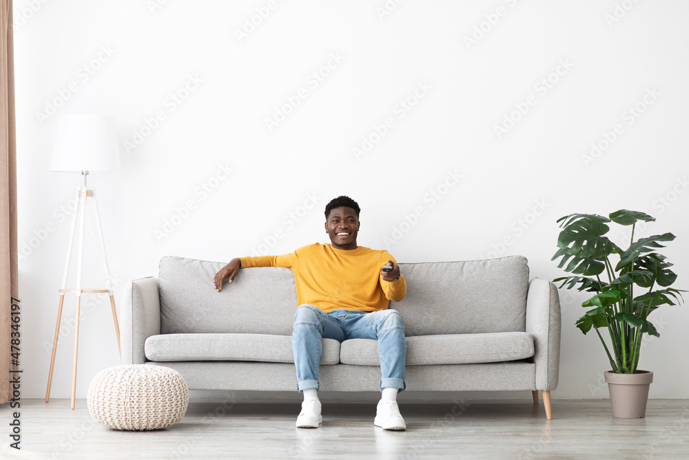 Joyful african american guy sitting on couch, turning on TV