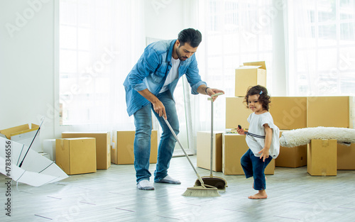 Handsome Caucasian father using broom sweep floor in living room at home or apartment, taking care daughter. Little naughty girl playing and walking around. Real Estate, Family moving house concept.
