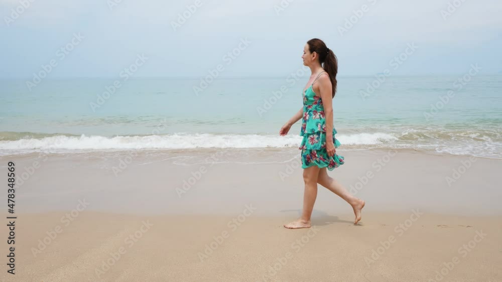 Relaxed tourist woman walk along edge of wet sand on sea shore, tracking camera motion. Hot and cloudy day at spring time in Thailand. Small waves break and wash sandy beach Relaxed tourist woman walk along edge of wet sand on sea shore, tracking camera motion. Hot and cloudy day at spring time in Thailand. Small waves break and wash sandy beach