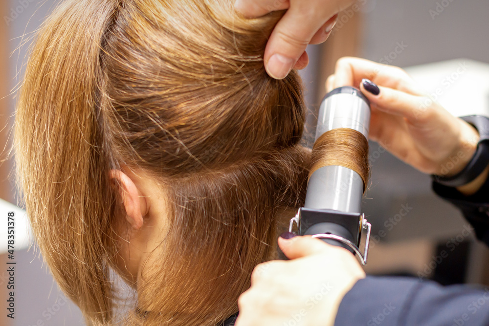 Fototapeta premium Hands of female hairstylist curls hair client with a curling iron in a hairdressing salon, close up