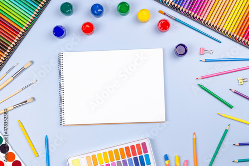 Creative art work supplies background. Watercolor, brushes, pencils, open blank notebook on blue table. Top view, flat lay