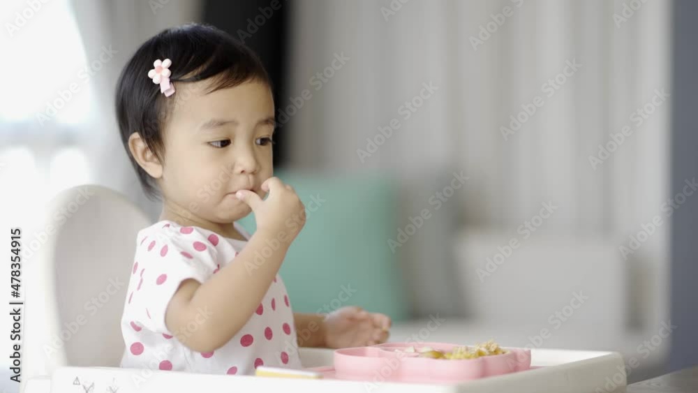 Asian cute little girl sitting on the white high chair and eating finger foods in the morning at home. Asian cute little girl sitting on the white high chair and eating finger foods in the morning at home.
