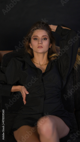 Portrait of a girl in a black dress. The girl is lying on the couch and looking at the camera. Thin figure. Black background.