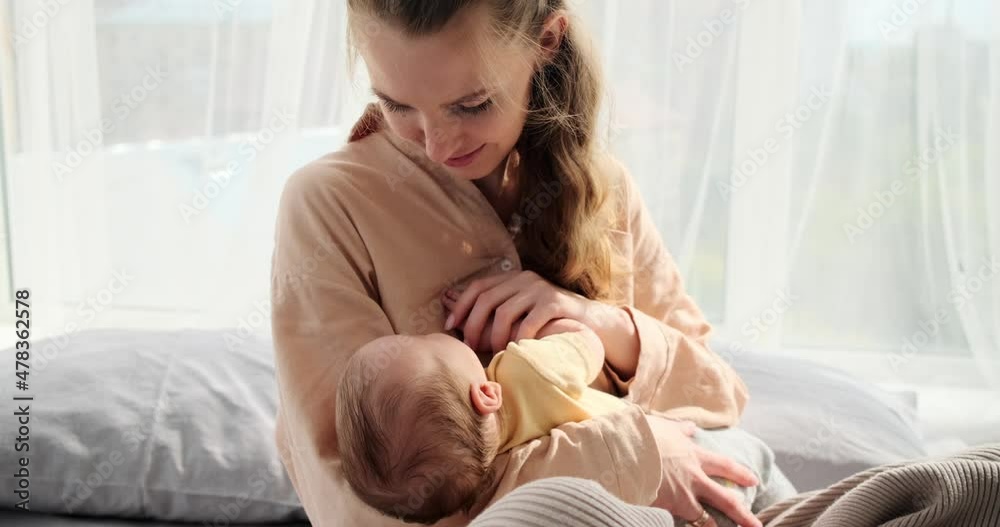 Young woman watching her baby son sleeping in arms sitting on bed at home Young woman watching her baby son sleeping in arms sitting on bed at home