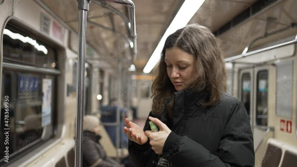 Young squeamish woman with dark wavy hair wearing warm jacket uses sanitizer on hands after holding handrail in underground carriage Young squeamish woman with dark wavy hair wearing warm jacket uses sanitizer on hands after holding handrail in underground carriage