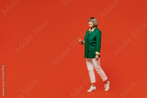 Full body side view fun elderly smiling happy woman 50s wearing green classic suit walk go point finger aside on workspace isolated on plain orange background studio People business lifestyle concept