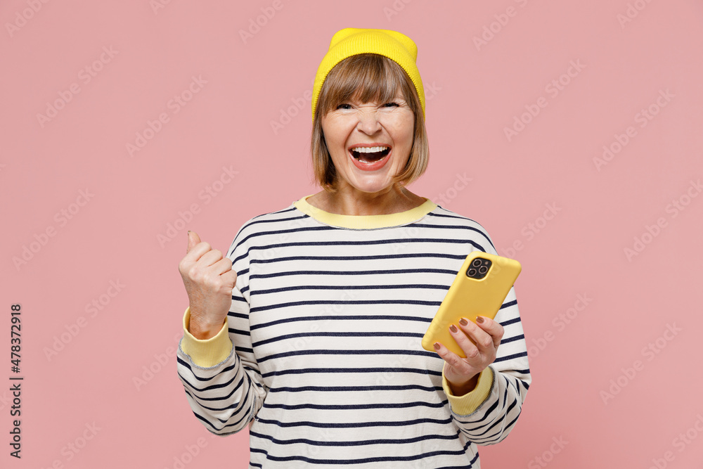 Elderly smiling happy fun woman 50s in striped shirt yellow hat hold using mobile cell phone do winner gesture isolated on plain pastel light pink background studio portrait. People lifestyle concept. Elderly smiling happy fun woman 50s in striped shirt yellow hat hold using mobile cell phone do winner gesture isolated on plain pastel light pink background studio portrait. People lifestyle concept.