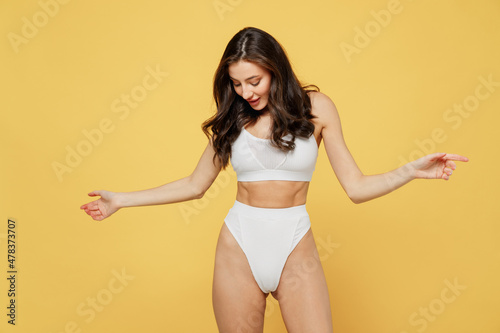 Smiling happy lovely attractive young brunette woman 20s wearing white underwear with perfect fit body standing posing look at waist hips shape isolated on plain yellow background studio portrait