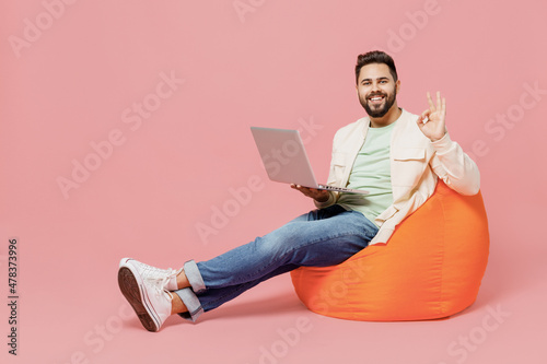 Full body young man in trendy jacket shirt sit in bag chair hold use work on laptop pc computer show ok okay gesture isolated on plain pastel light pink background studio. People lifestyle concept