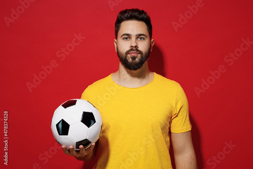 Young bearded man football fan in yellow t-shirt cheer up support favorite team hold in hand soccer ball isolated on plain dark red background studio portrait. People sport leisure lifestyle concept. © ViDi Studio