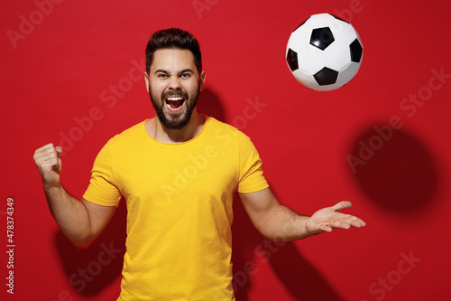 Exultant fun young bearded man football fan in yellow t-shirt cheer up support favorite team tossed soccer ball celebrate clenching fists say yes isolated on plain dark red background studio portrait. photo