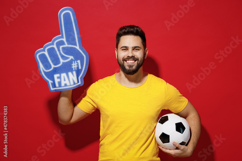 Blithesome young bearded man football fan in yellow t-shirt cheer up support favorite team look camera hold soccer ball fan foam glove finger up isolated on plain dark red background studio portrait. photo